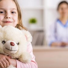 Niña Mirando Al Frente, En El Fondo Se Ve Una Persona En Un Mostrador En Un Centro De Asistencia De Salud